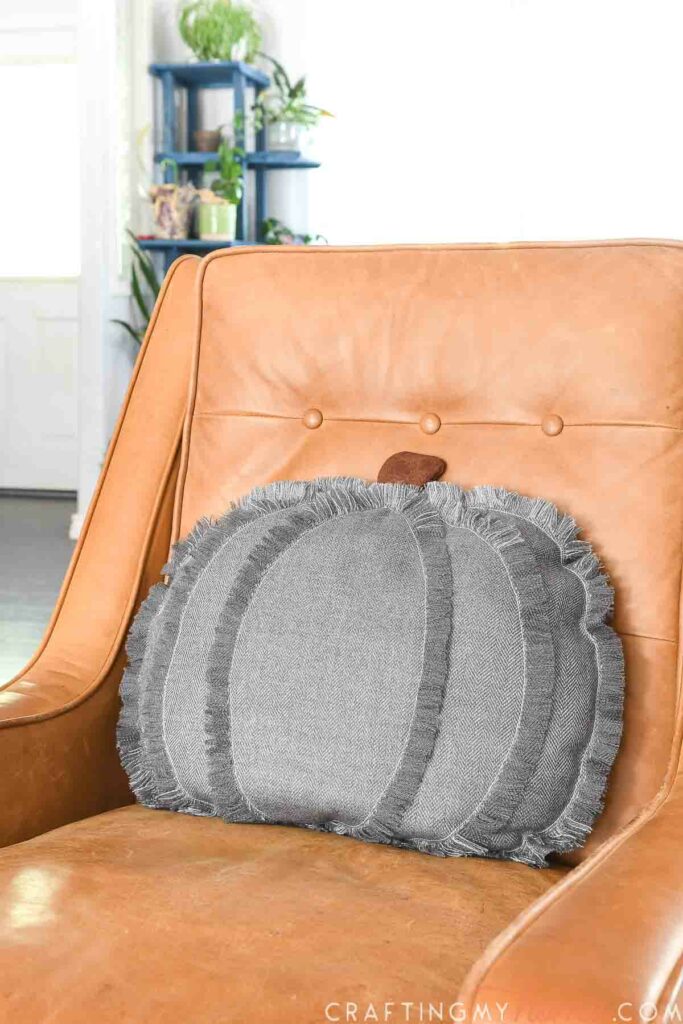 Modern pumpkin pillow with frayed edges and faux leather stem on a camel colored chair. 