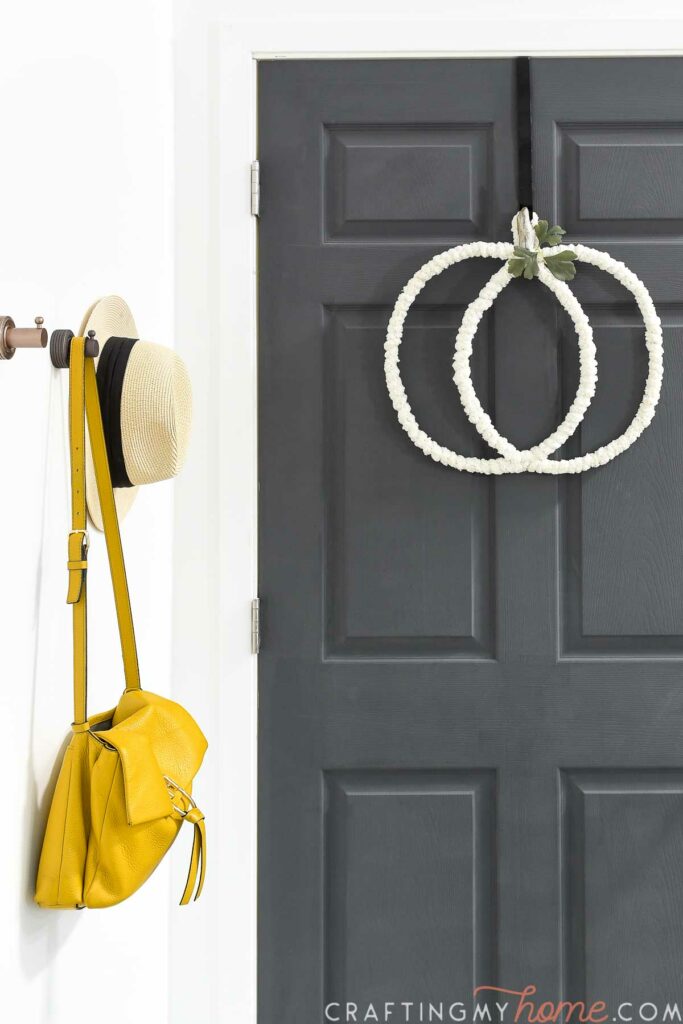 Black painted door with wire pumpkin wreath on the front and hooks next to it with a purse and hat on them. 