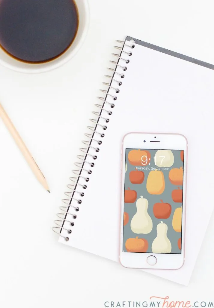 Smartphone sitting on a notebook next to a cup of coffee with free digital background with pumpkin design on the home screen. 