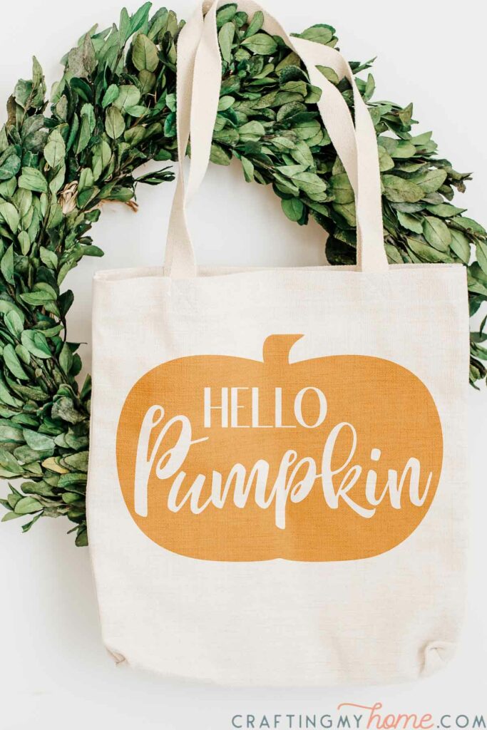 Canvas bag with Hello Pumpkin SVG design stenciled onto the front hanging in front of a wreath. 