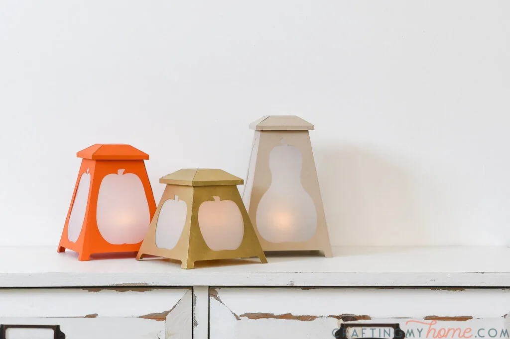Three tapered paper lanterns with pumpkin and gourd shapes cut out of the sides. 
