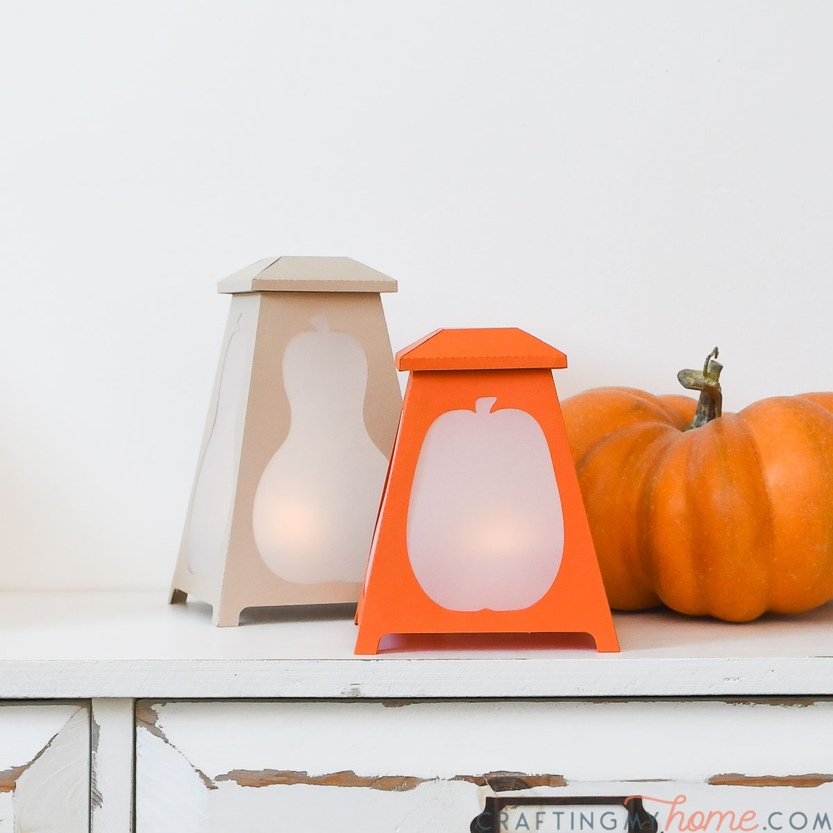 Two tapered lanterns with pumpkin cutouts sitting on a shelf next to a decorative pumpkin.